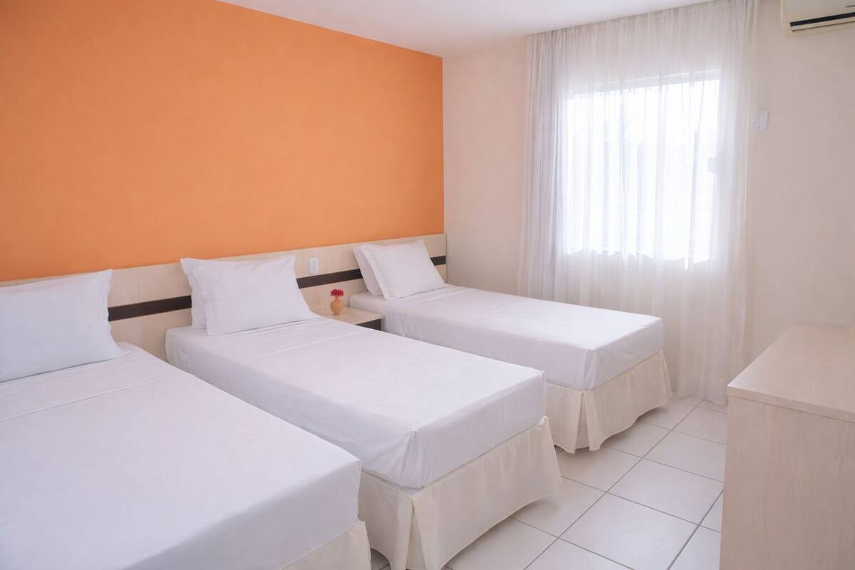Habitación de hotel con dos camas individuales, una mesita de noche pequeña con un florero, una ventana con cortinas traslúcidas y un equipo de aire acondicionado blanco en la pared.