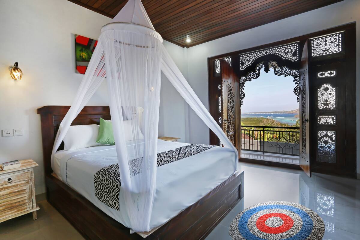 Ein Schlafzimmer mit einem Bett unter einem weißen Baldachin, einem Nachttisch, einem dekorativen Teppich und Blick durch eine Holztür.