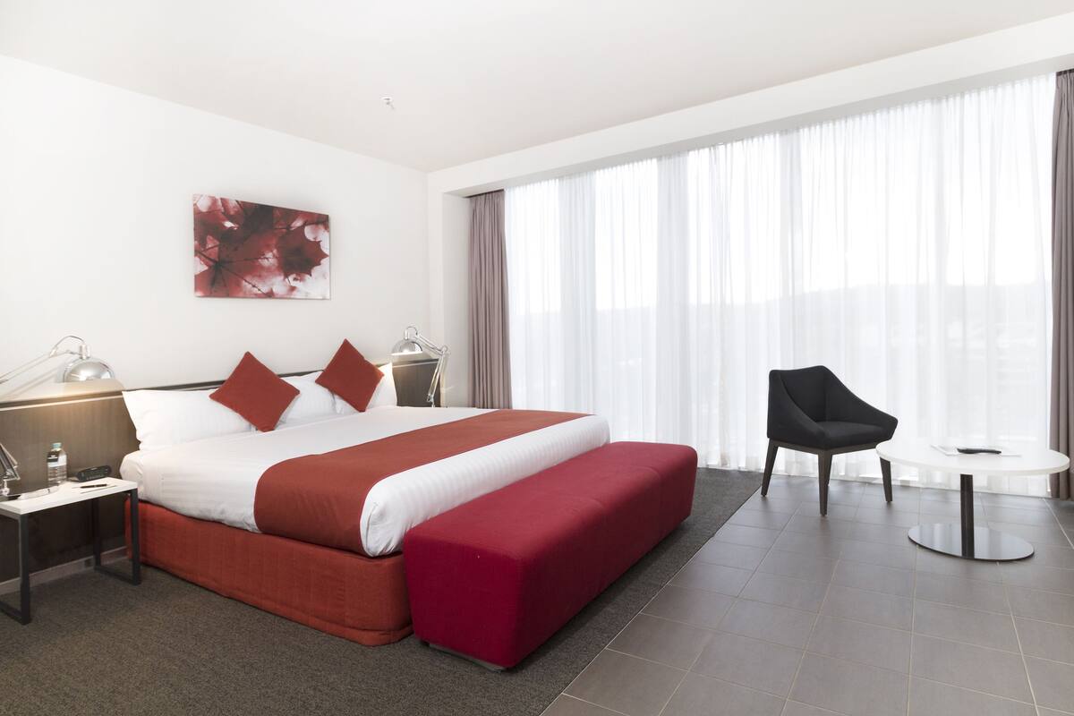 Habitación de hotel con una cama grande, una mesita de noche con lámpara, una silla, una pequeña mesa y un ventanal con cortinas.