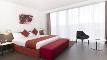 A hotel room with a large bed, a bedside table with a lamp, a chair, a small table, and a large window with curtains.