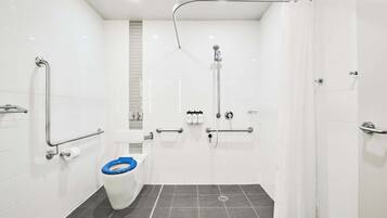Uma casa de banho moderna com azulejos brancos, chuveiro com teto curvo, sanita com assento azul e barras de apoio.