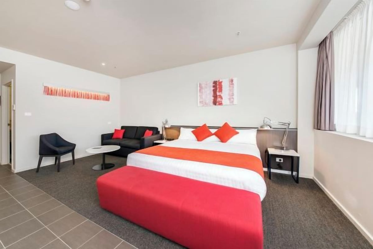 A hotel room with a large bed, a sofa, a chair, a small table, and a lamp.