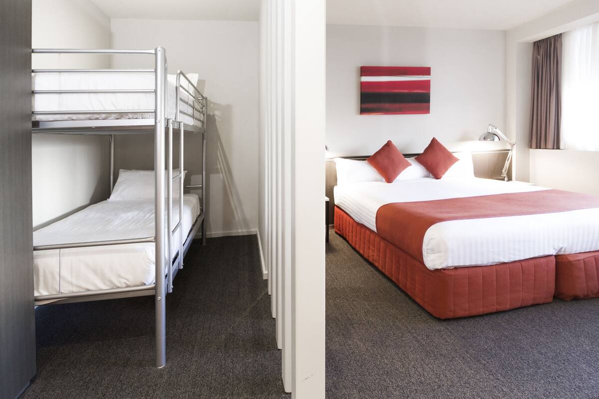 Una habitación con una cama litera y una cama individual, ambas con ropa de cama blanca. Hay una lámpara en una mesita de noche y un cuadro en la pared.