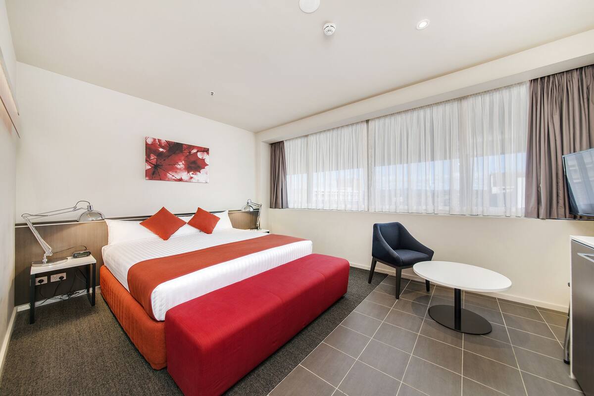 Habitación de hotel con cama, mesita de noche, lámpara, silla, mesita redonda y televisor.