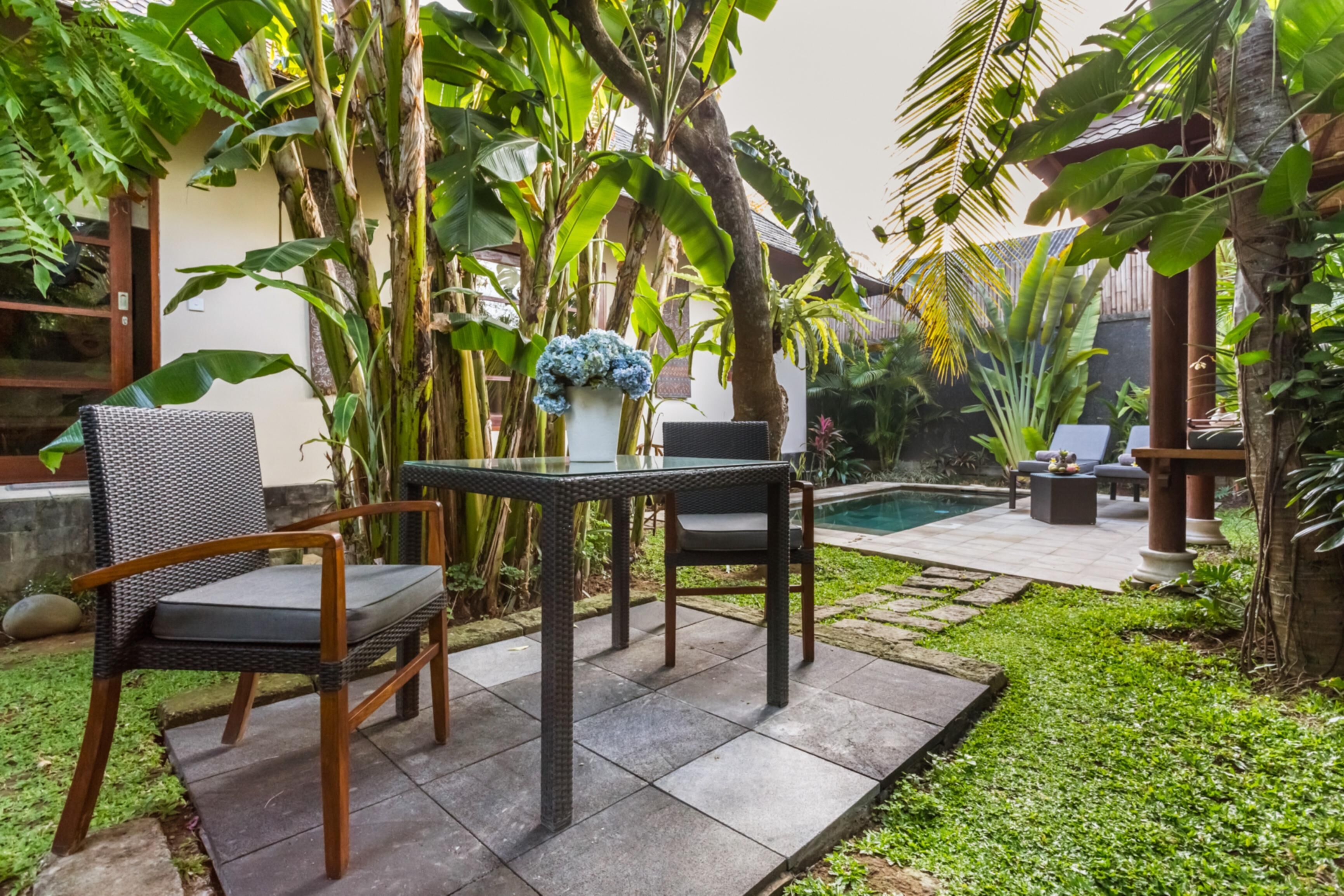 Un patio al aire libre con una mesa y sillas, rodeado de abundante follaje y una piscina.