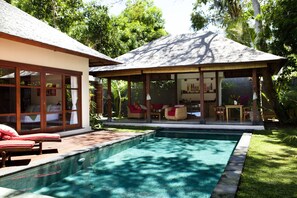 Un espace aménagé au bord de la piscine, comprenant un coin salon couvert, une pièce fermée par des baies vitrées et une pelouse bien entretenue.