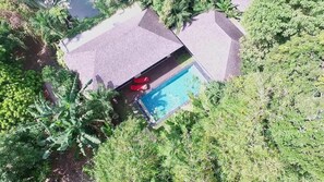 Vue aérienne d’une propriété dotée d’une piscine entourée d’une végétation luxuriante et d’un espace abrité avec des chaises longues rouges.