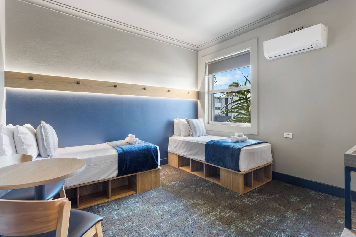 Habitación de hotel pequeña con dos camas individuales, una mesita y una ventana que da a palmeras.