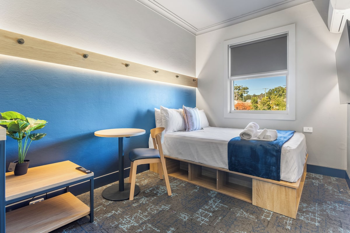 Una habitación pequeña con cama, una mesita, una silla y una planta en maceta.