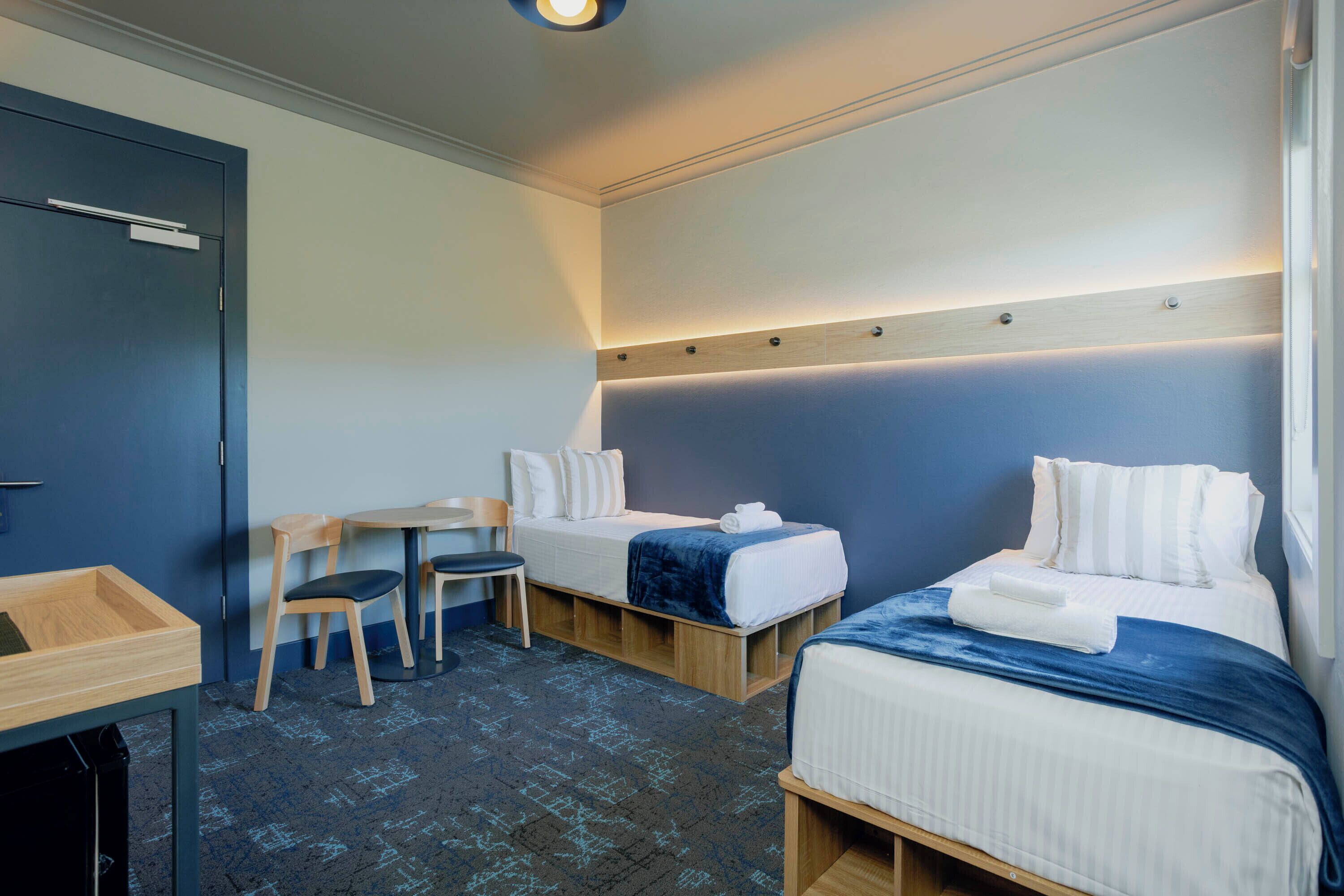 A hotel room with two single beds, a small desk, a chair, and a wooden cabinet.
