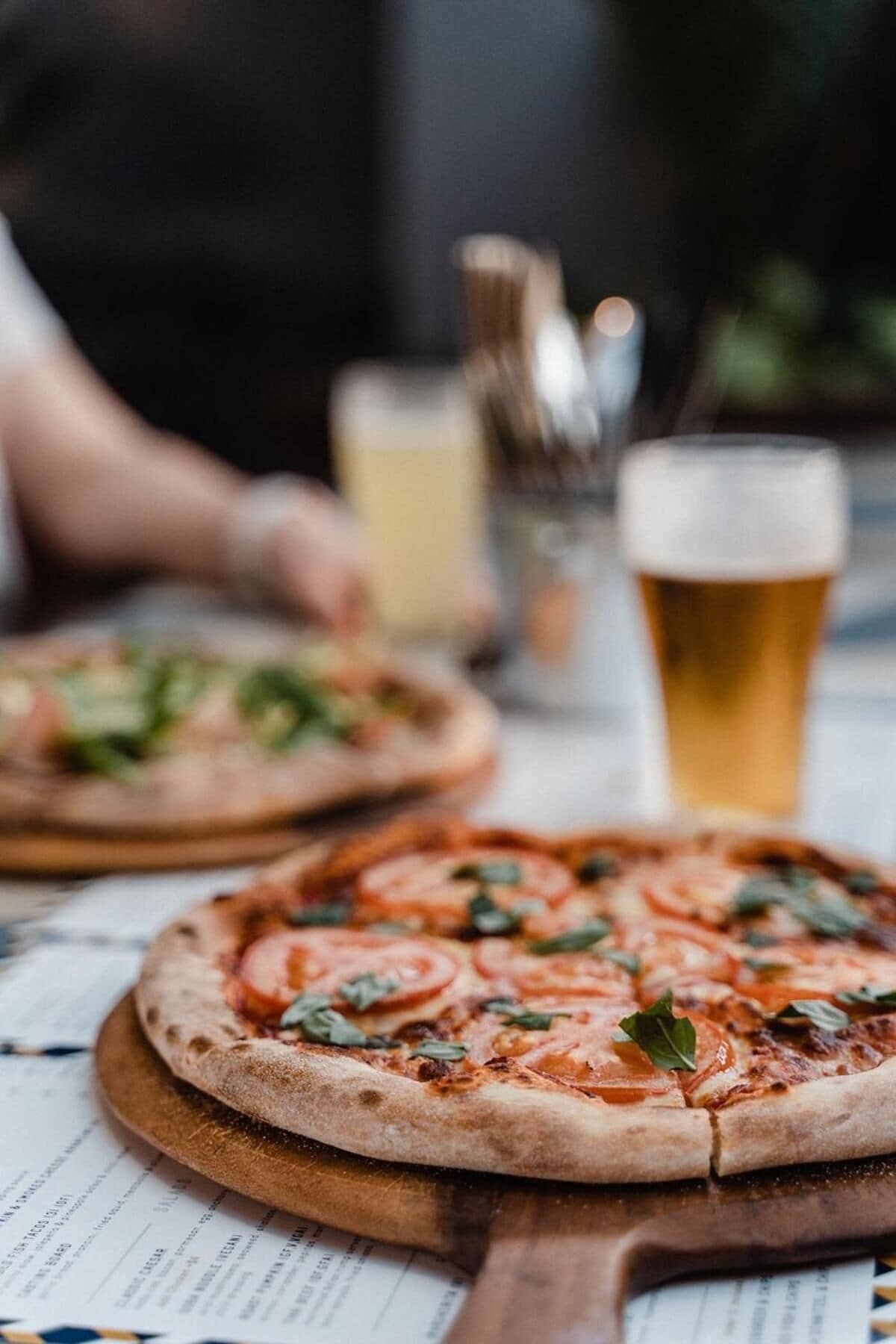 Two pizzas on wooden boards with a menu underneath, and a glass of beer in the background.