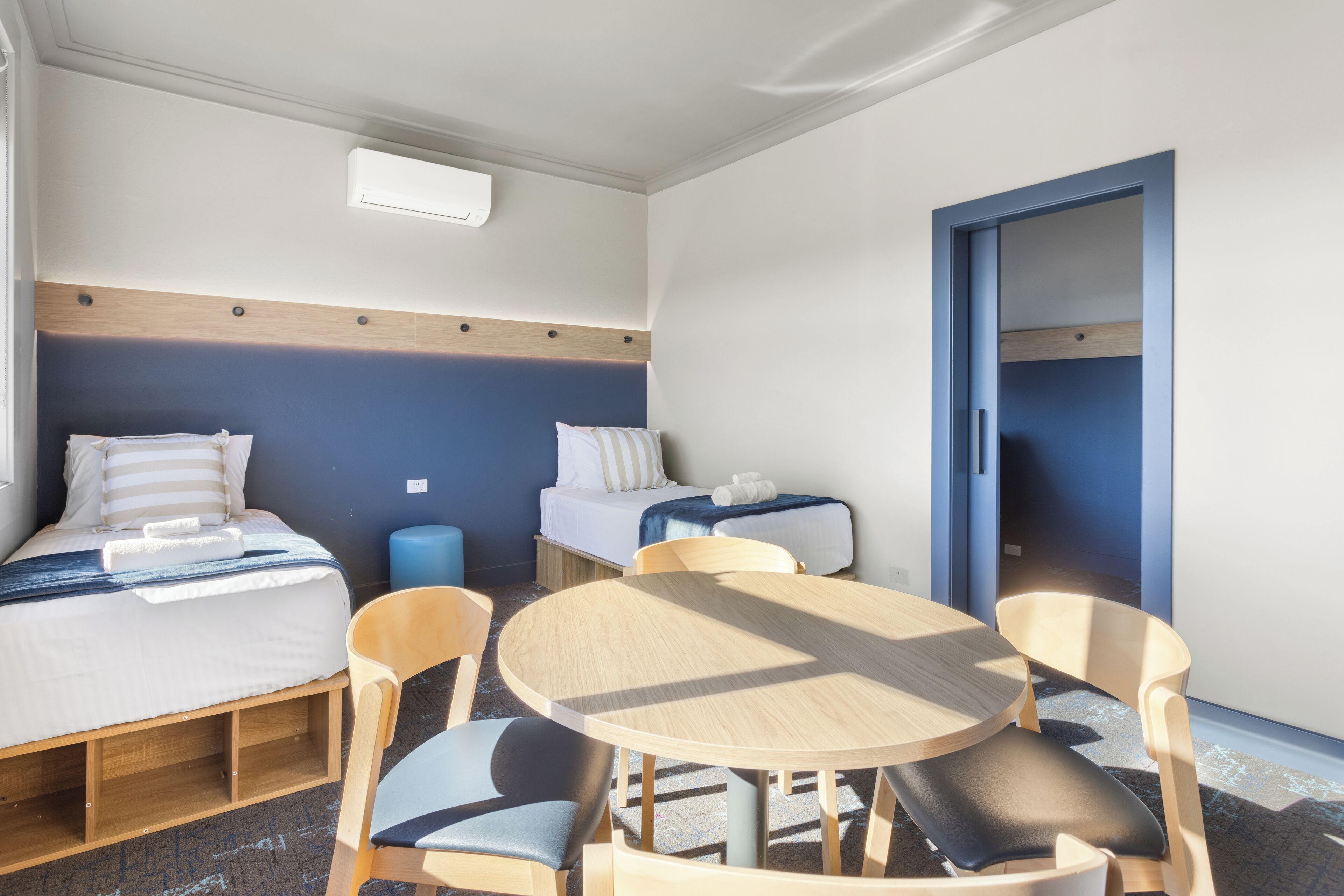 Quarto de hotel com duas camas, uma mesa redonda de madeira e quatro cadeiras.