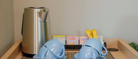 A kitchen area with a stainless steel kettle, blue plates, and a tray with packaged food items.