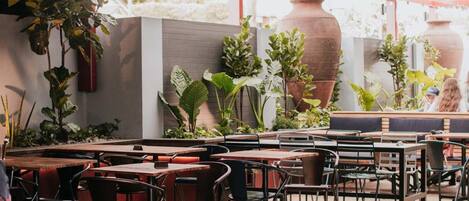 An outdoor cafe with tables and chairs, potted plants, and a red ceiling.