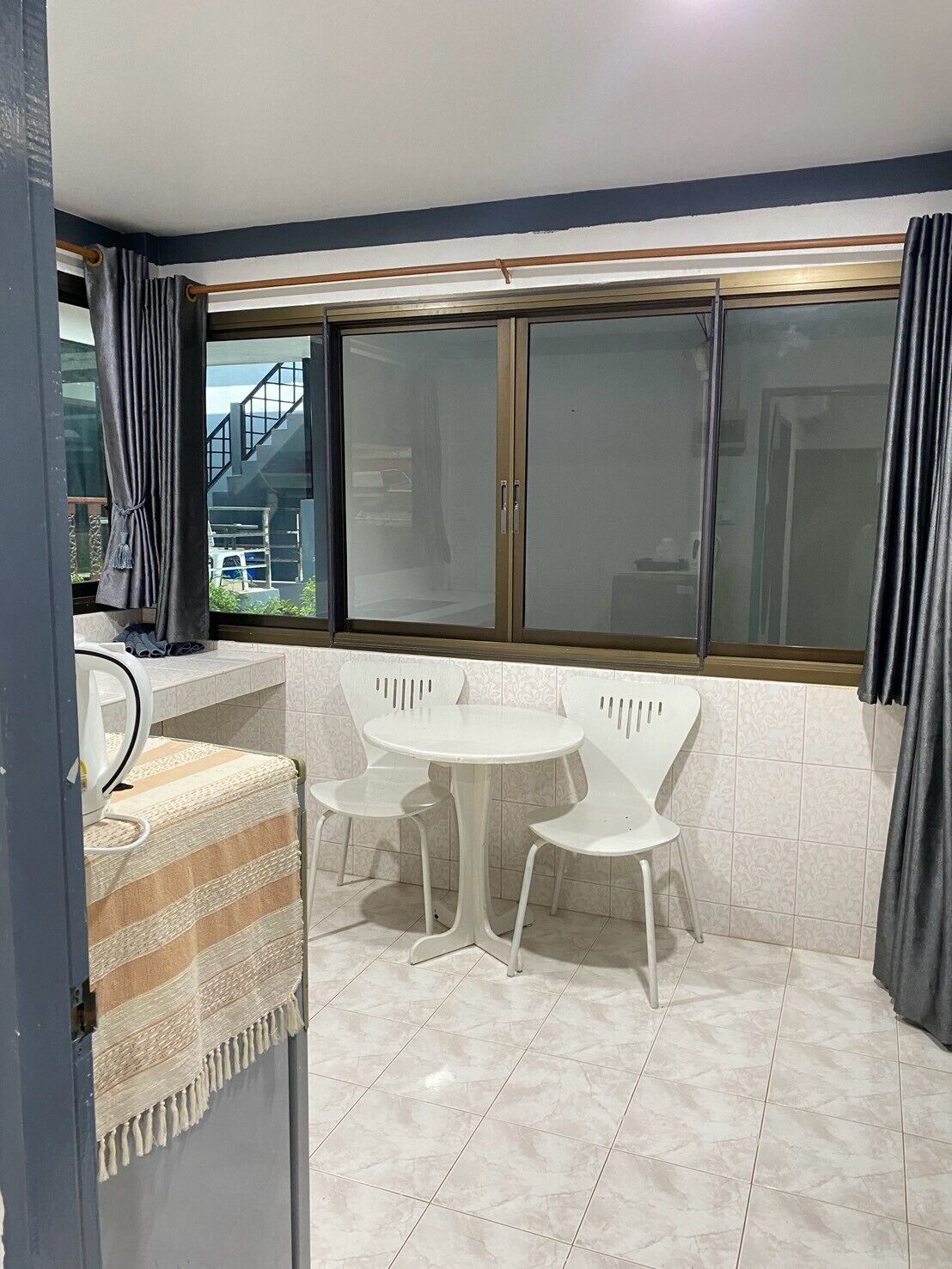 Un pequeño comedor con una mesa redonda y cuatro sillas, una ventana con cortinas y un suelo de baldosas.