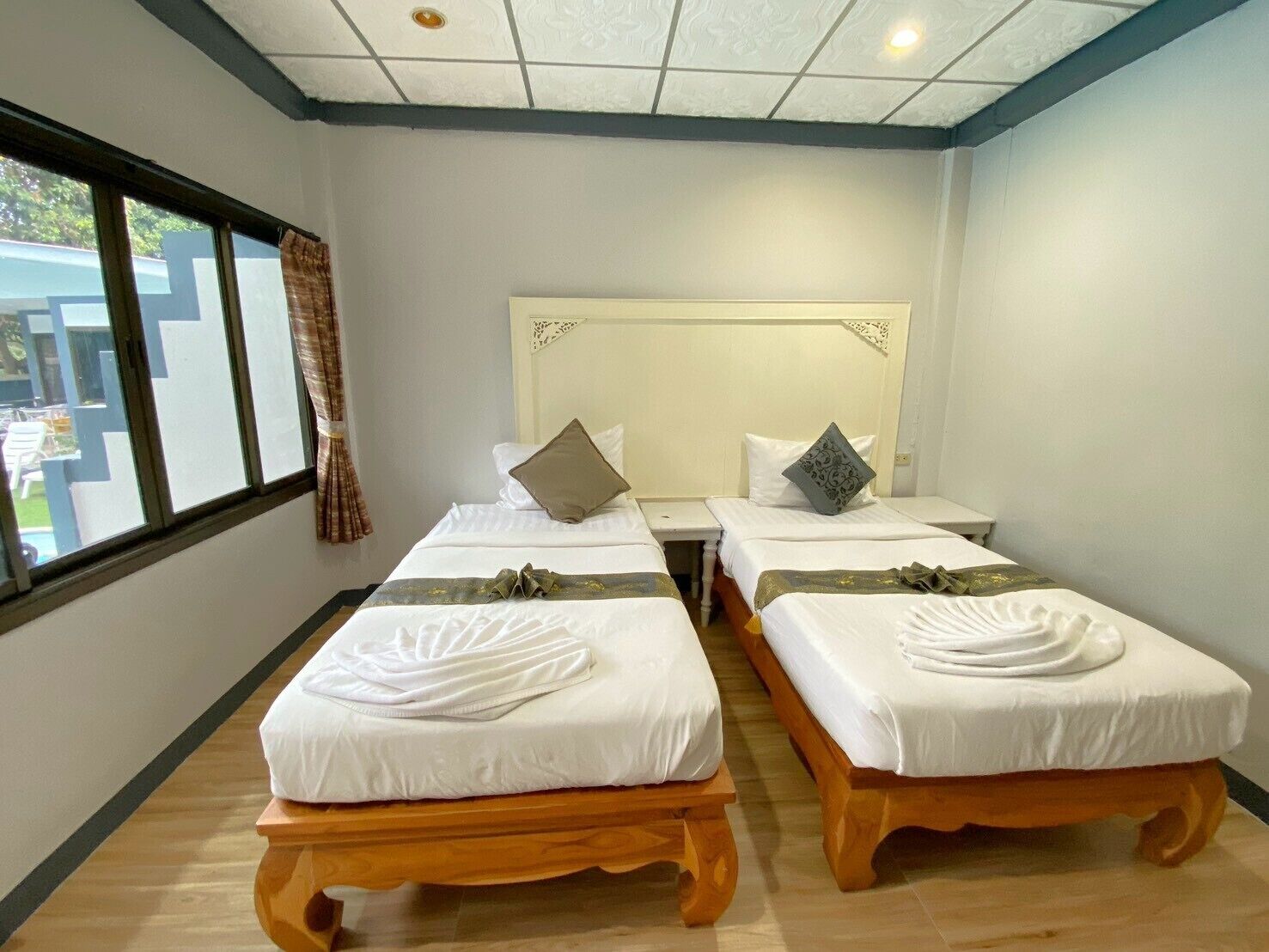 A hotel room with two single beds, a wooden floor, a window with curtains, and a view of the outside.
