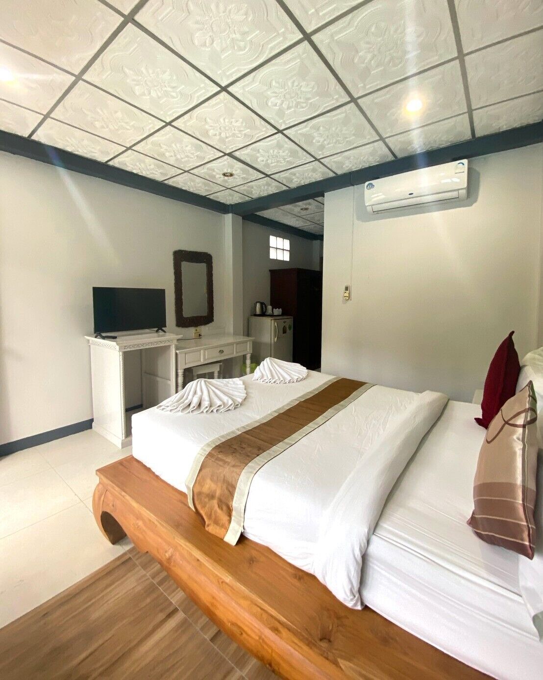 A hotel room with a bed, a wooden bench, a desk with a television, a mirror, and an air conditioning unit.