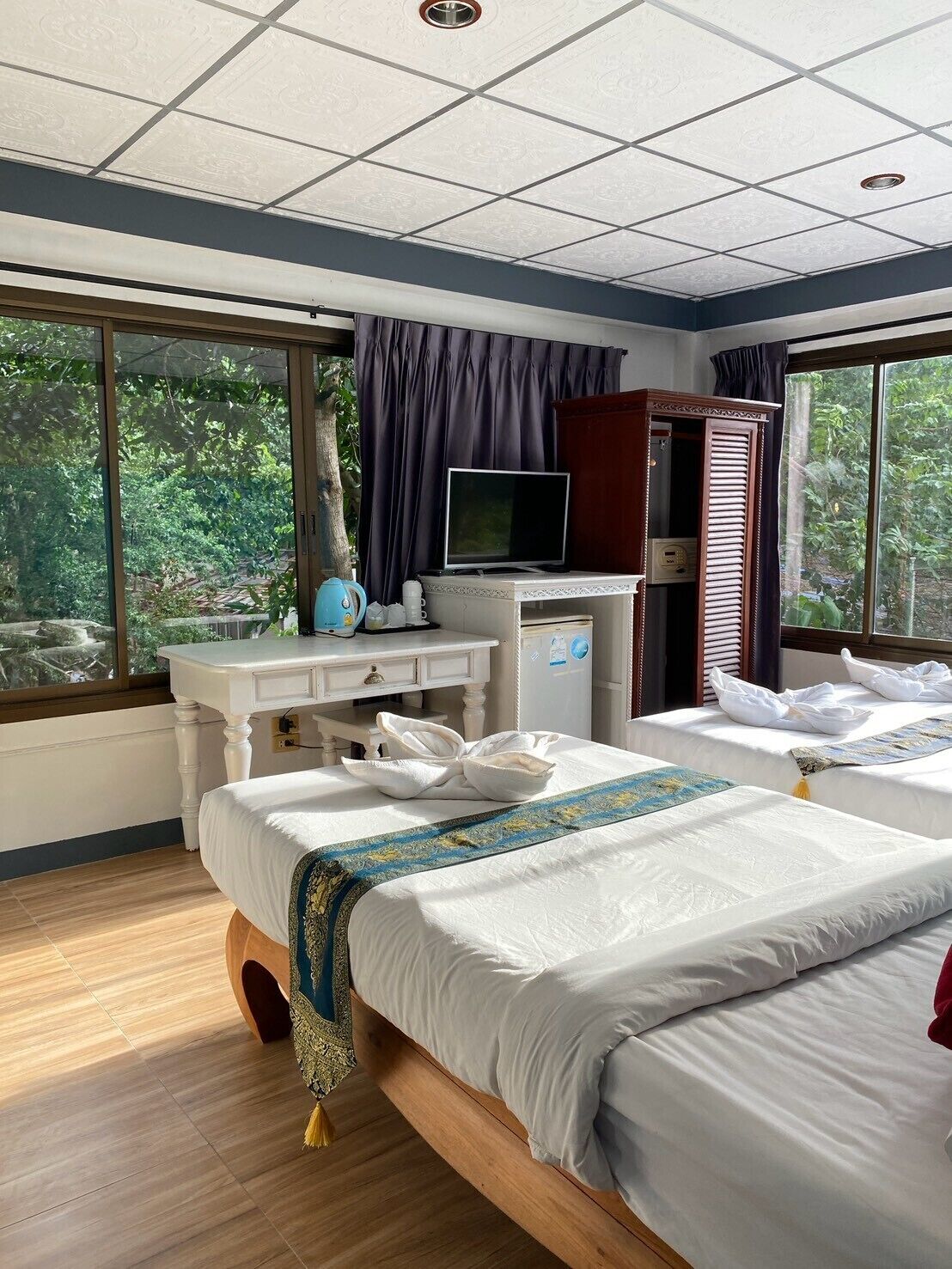 Quarto de hotel com cama, escrivaninha com televisão e janelas amplas.