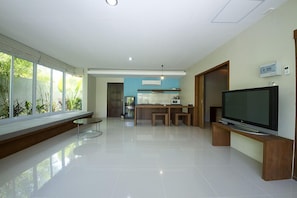 A modern living room with a large TV, a dining area, and a kitchen in the background. - The Living Pool Villas (Koh Samui)