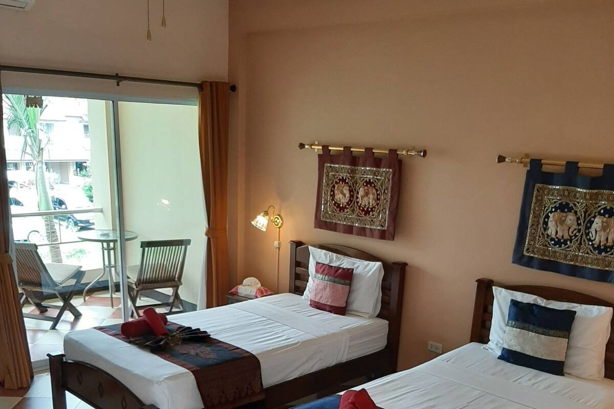 Quarto de hotel com duas camas, um ventilador de teto, janela com cortinas, luminária na parede e vista para o exterior.