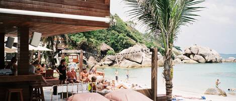 Zona de piscina con sillas de descanso y un bar, con vista a una playa con rocas y palmeras.