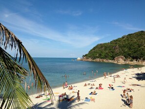Una playa con gente tomando el sol y nadando, una orilla arenosa y una colina con árboles al fondo.
