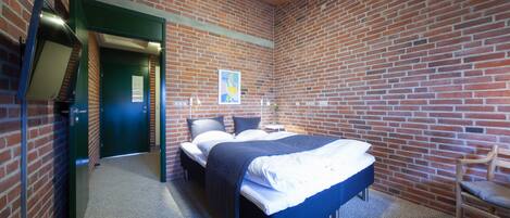 Una habitación con pared de ladrillo, una cama con ropa de cama blanca y azul, una mesita con una lámpara y una silla.