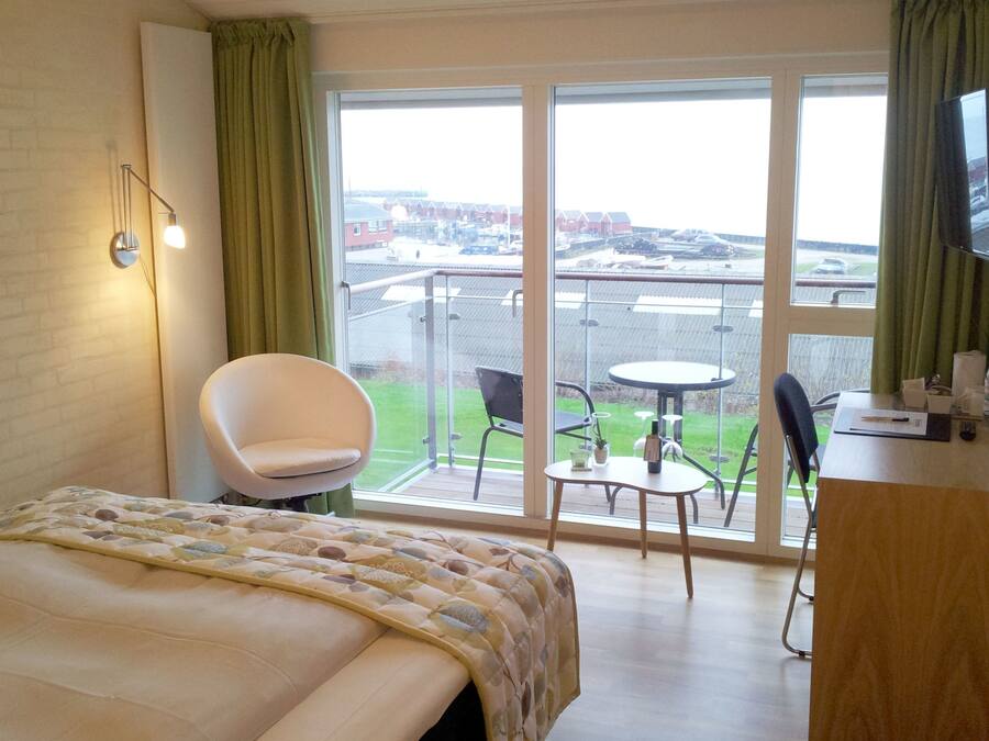 Habitación de hotel con cama, una silla, una mesita, televisión y balcón con vista a la ciudad.