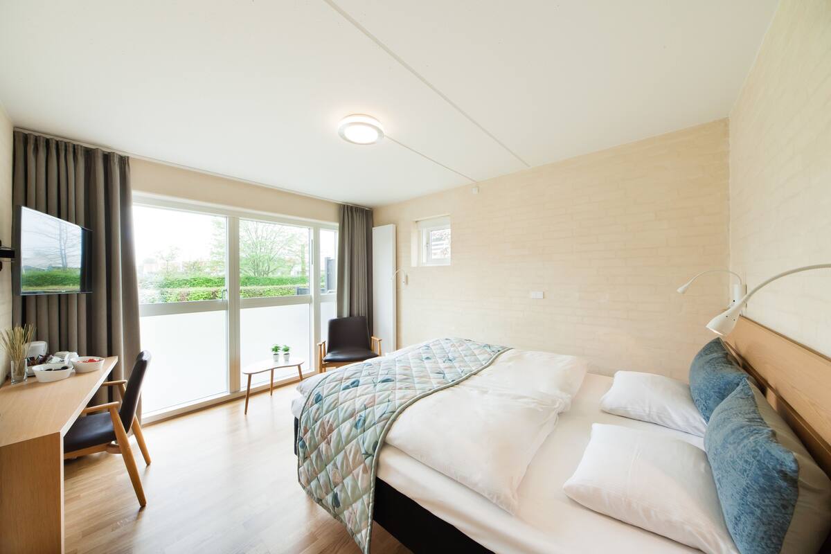 Habitación de hotel con cama, escritorio, silla, televisión y ventana con vistas a zonas verdes.