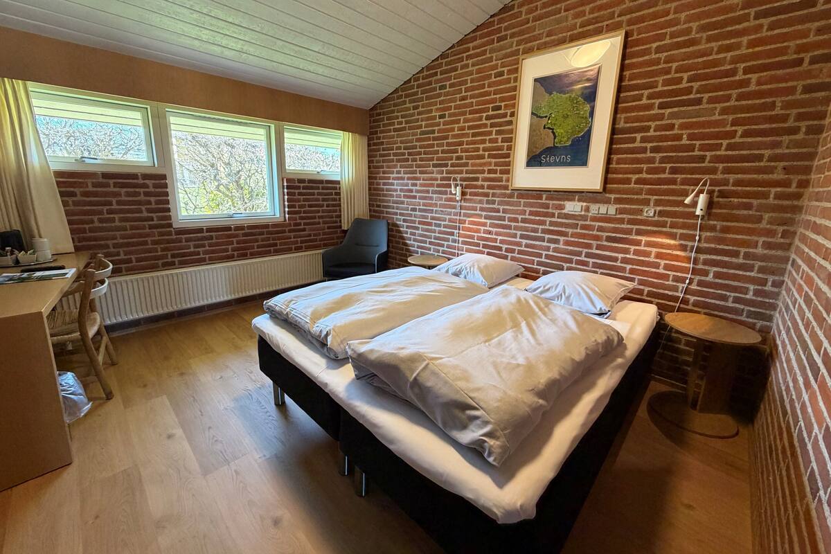 Una habitación con pared de ladrillo, cama con ropa de cama blanca, escritorio con silla y un mapa en la pared.