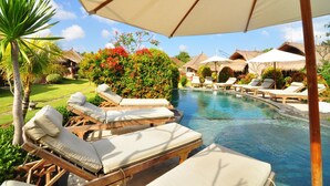 A poolside area with lounge chairs and umbrellas, surrounded by lush greenery and a clear sky.