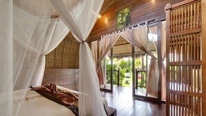 A room with a bed, a wooden floor, a window with a view of greenery, and a wooden partition.