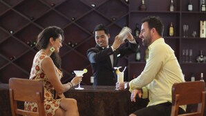 A bar setting with a bartender preparing drinks. There are three people seated at the bar, and shelves with bottles in the background. - Sokhalay Angkor Residence and Spa (Siem Reap)