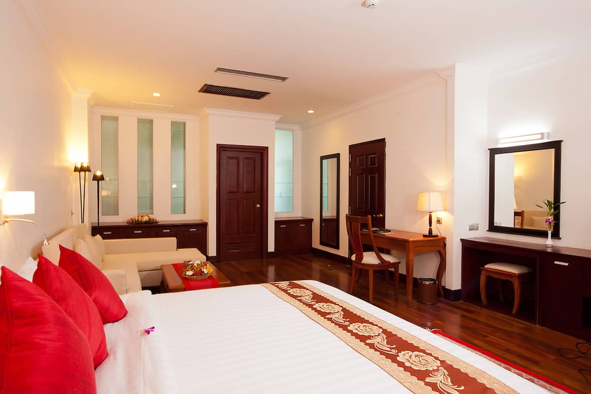 A hotel room with a large bed, a desk, a chair, a small table, a mirror, and a lamp.