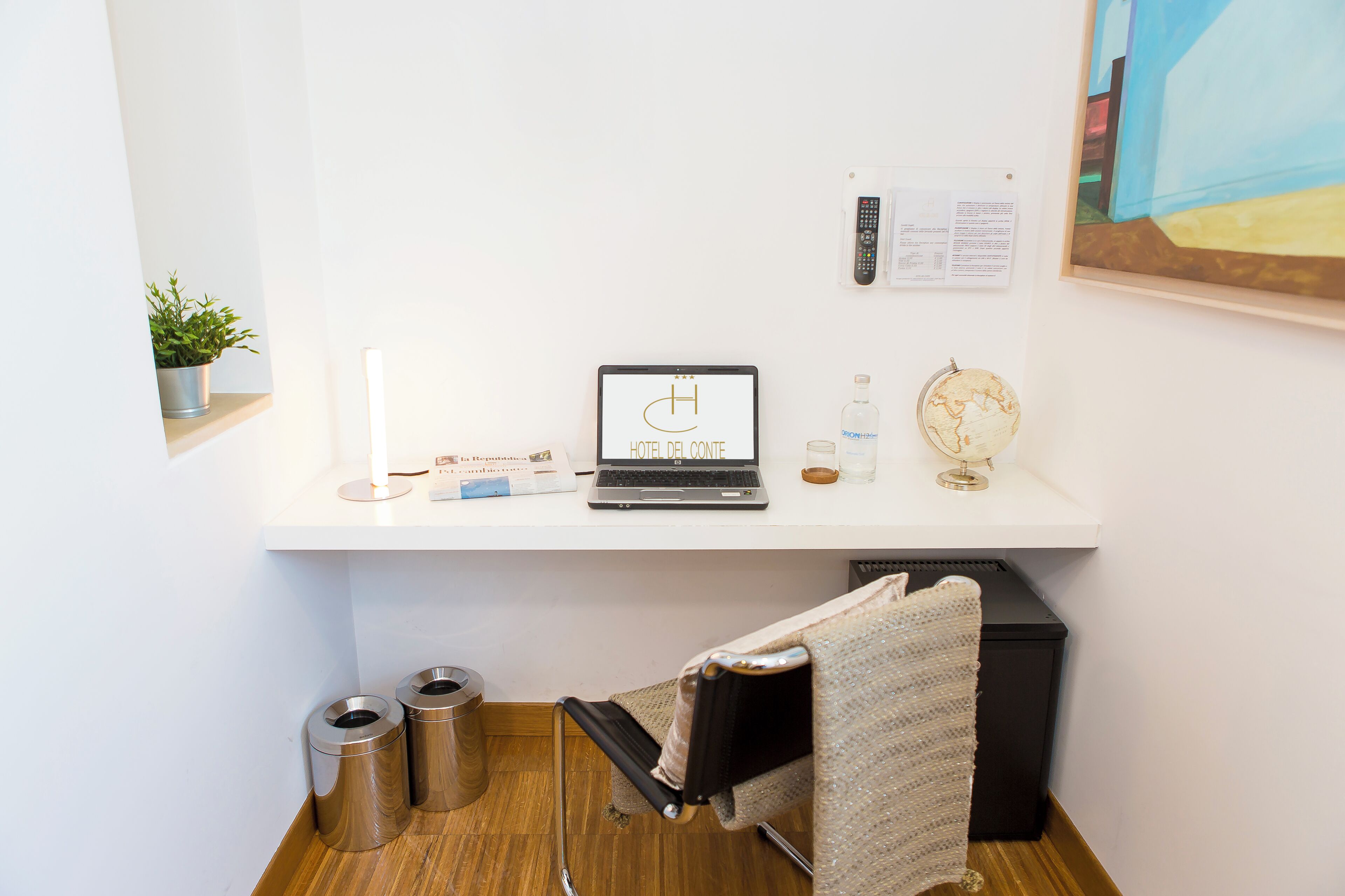 Een nette, moderne hotelkamer met een bureau, laptop, plant, wereldbol en een bord met een welkomstboodschap.
