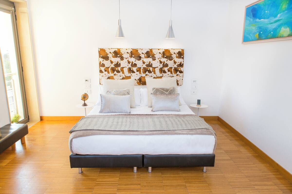 A modern bedroom with a large bed, two bedside tables, a headboard with a unique pattern, and a painting on the wall.
