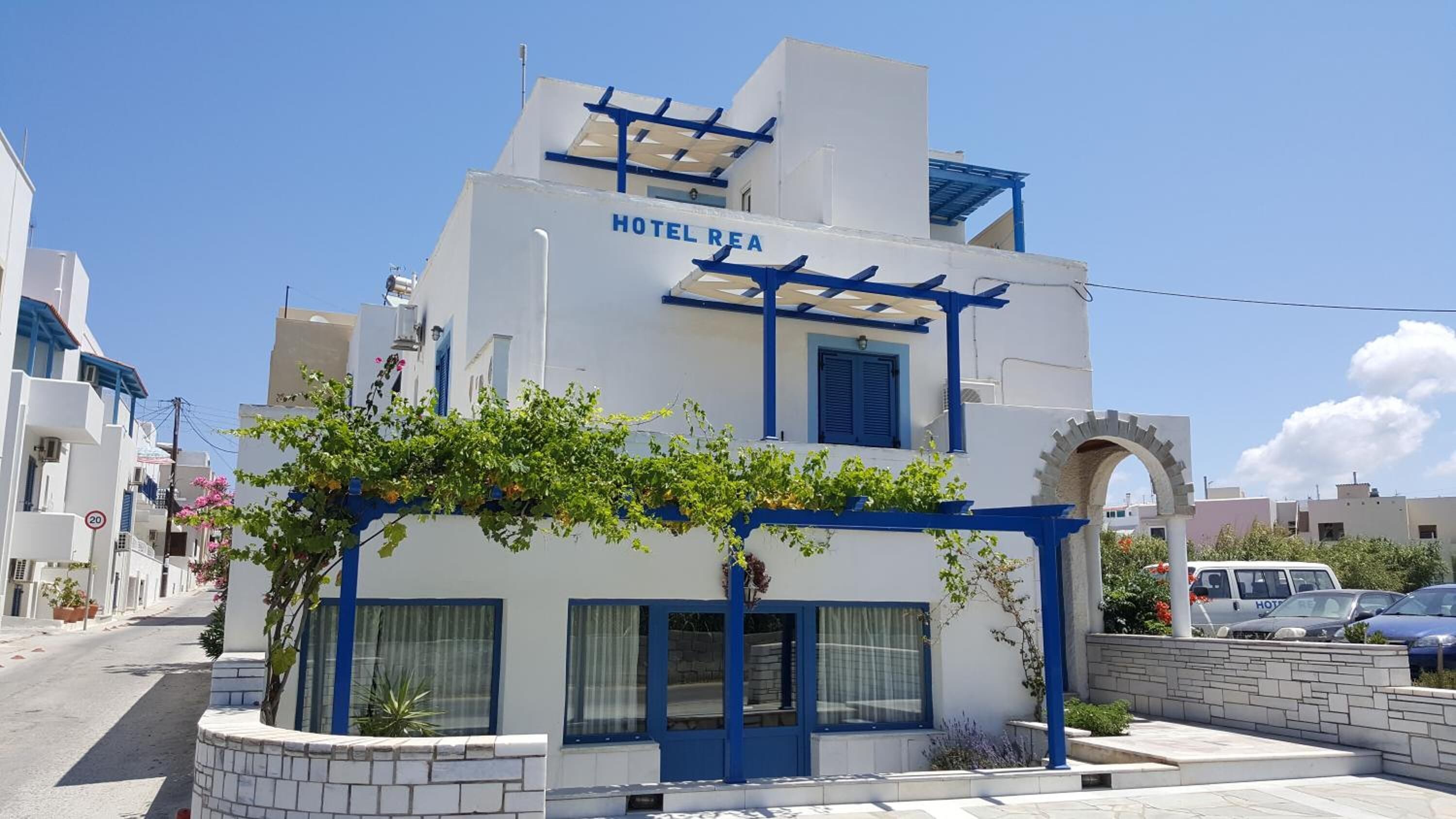 A white building with blue accents, labeled "HOTEL REA," featuring a central entrance with a pergola and a stone wall.