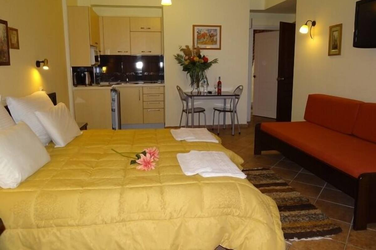 Habitación de hotel con cama, sofá, mesa de comedor con sillas, zona de cocina y televisor.