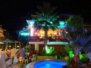 A pool area with a dining terrace, palm trees, and a building with illuminated signage.