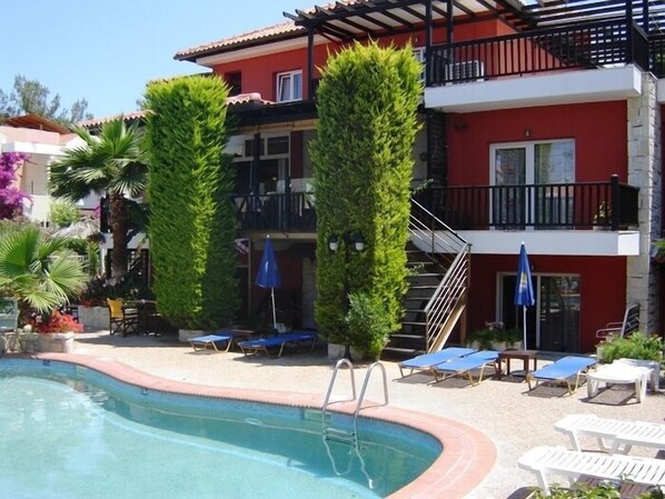 A hotel with a swimming pool, sun loungers, and a red building with balconies.