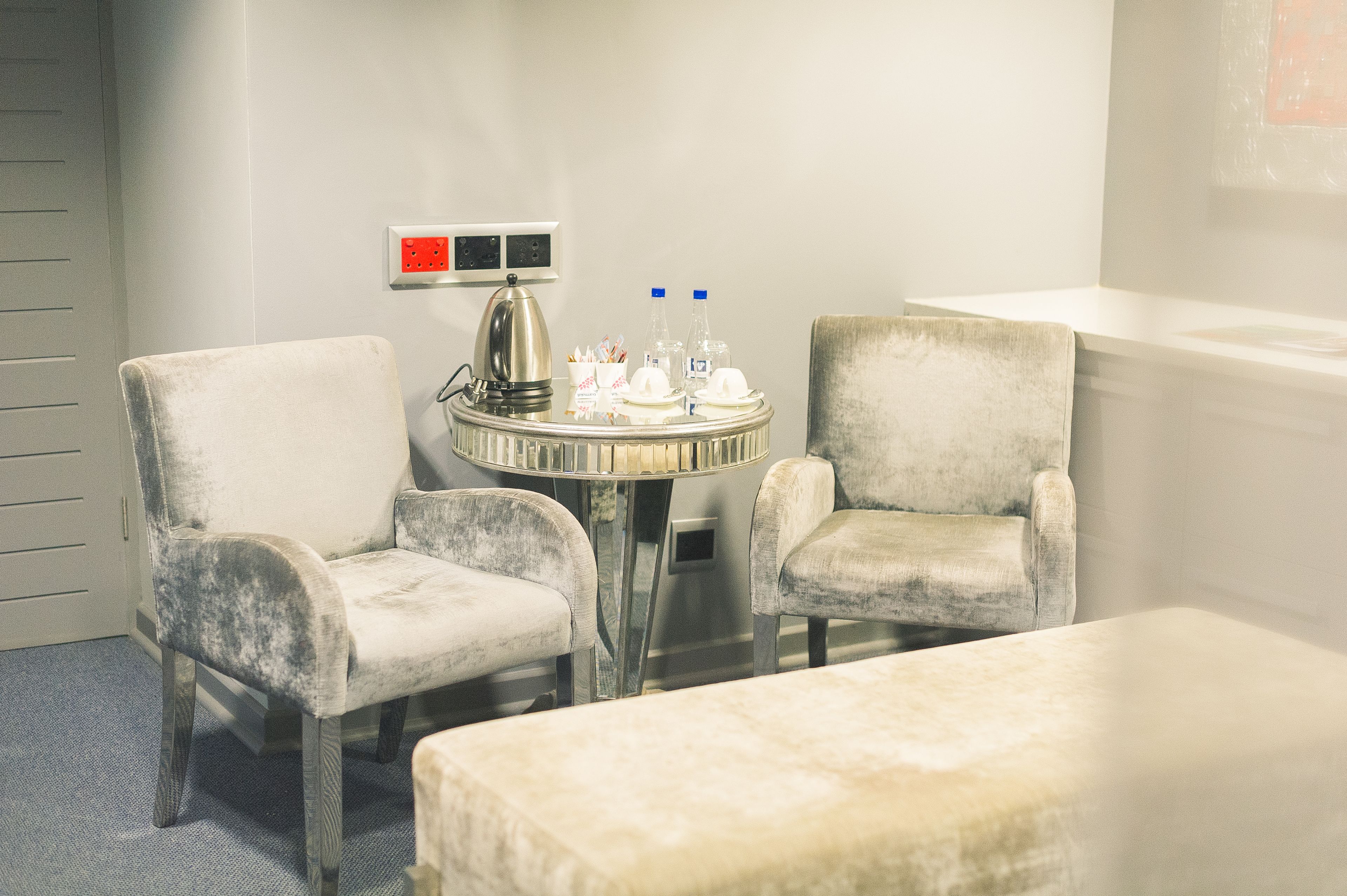 Habitación de hotel con dos sillones, una mesita redonda con hervidor, tazas y botellas, y una mesita de noche blanca.