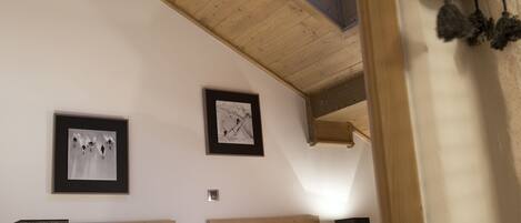 Ein Schlafzimmer mit schrägem Dach, Holzbalken, einem Oberlicht, einem Bett mit roter Decke, zwei gerahmten Bildern, einem Teddybären und Nachttischlampen.