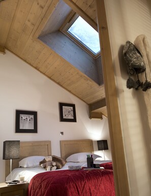Un dormitorio con techo inclinado, vigas de madera, tragaluces, cama con manta roja, dos cuadros enmarcados, un osito de peluche y lámparas de noche.