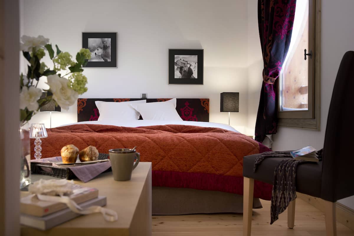 Ein ordentlich eingerichtetes Hotelzimmer mit Bett, Nachttisch mit Lampe, Stuhl und einem Fenster mit Vorhängen.