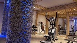 Ein Fitnessraum mit Trainingsgeräten, Holzbalken und einer säulenförmigen Struktur mit Kies.