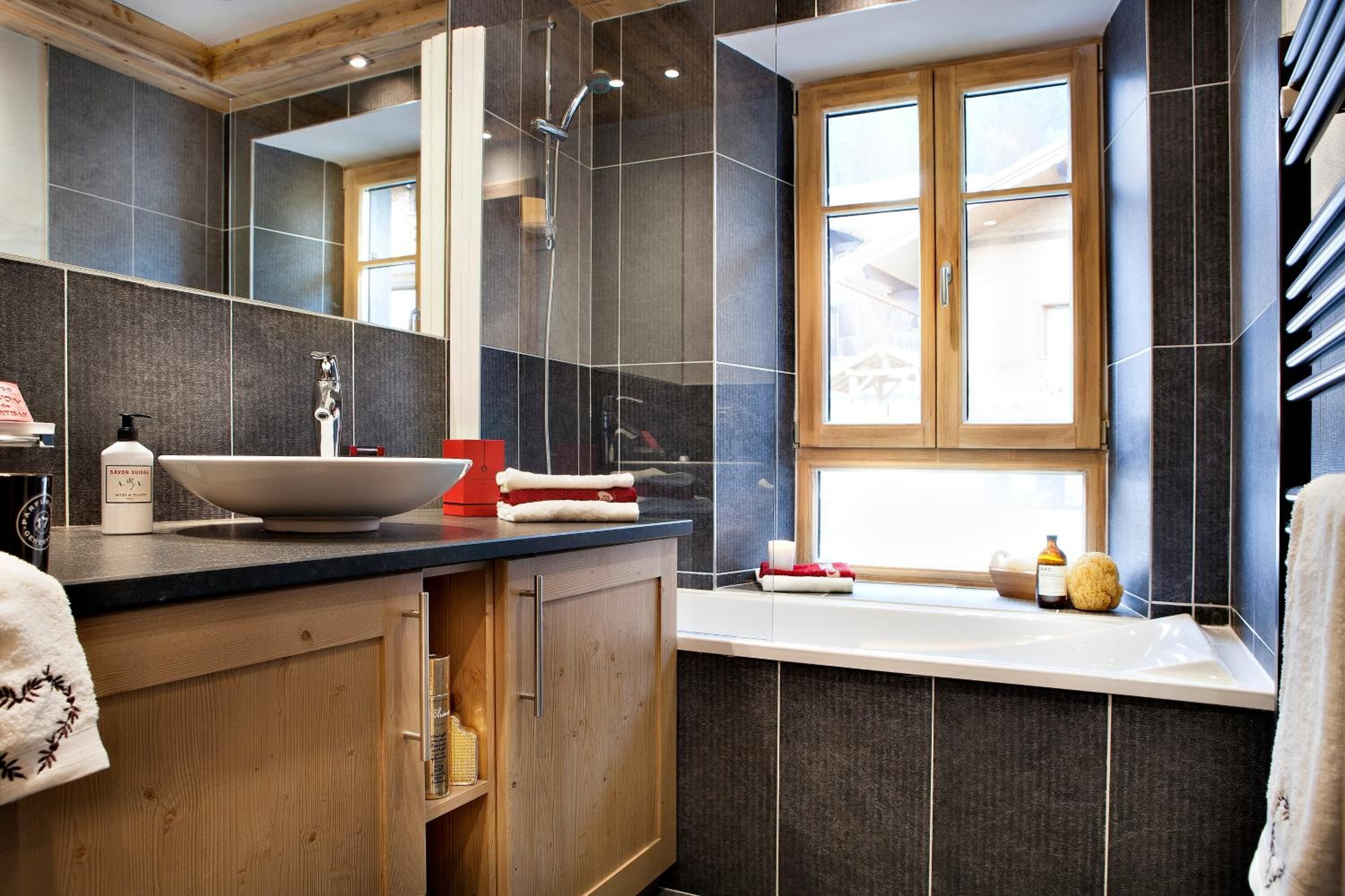 Une salle de bain moderne avec un grand miroir, des carreaux foncés, un meuble-lavabo en bois, un lavabo blanc et une fenêtre.