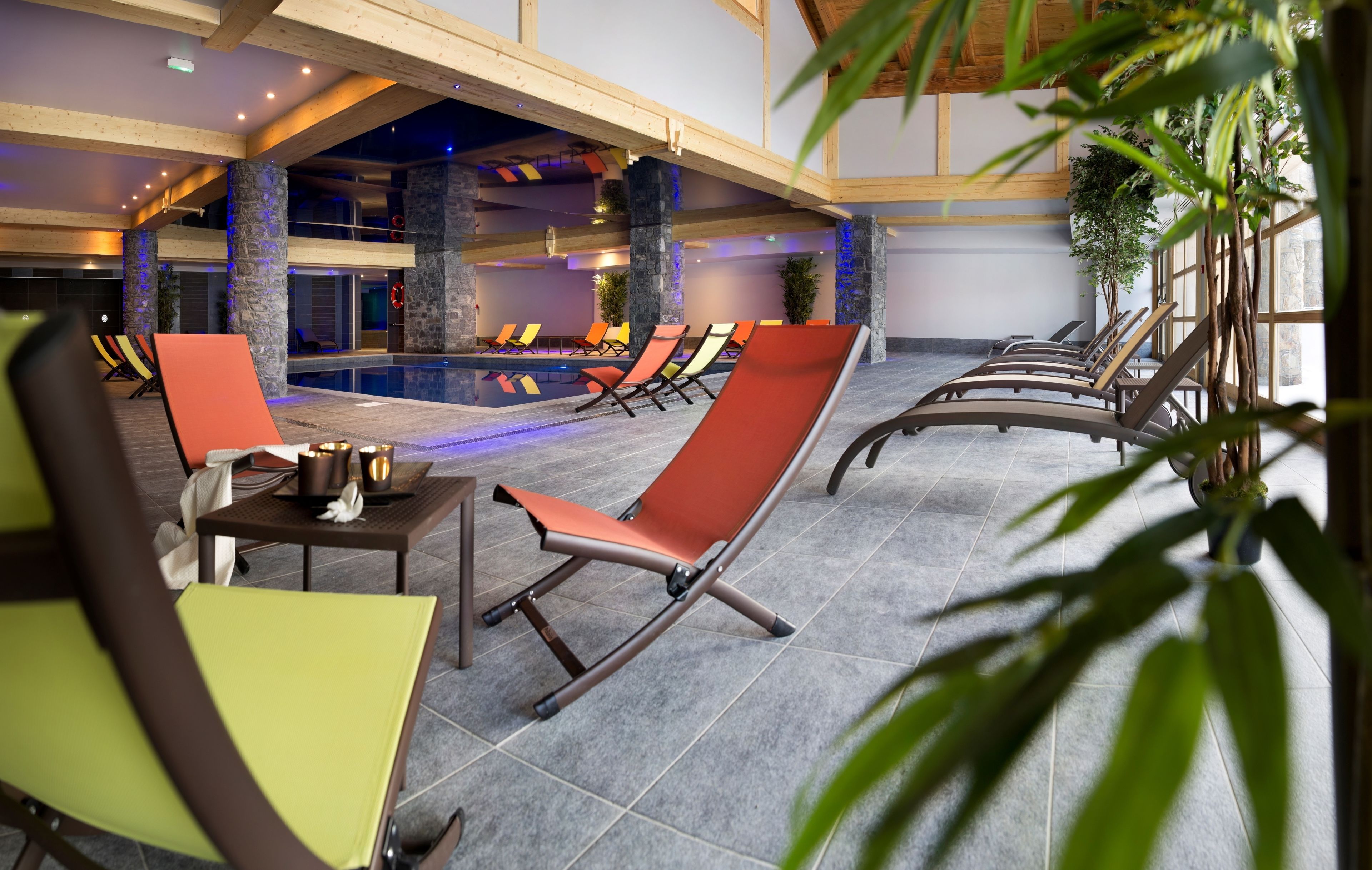 Uma área à beira da piscina com espreguiçadeiras e uma mesa, piscina interna e pilares de pedra.