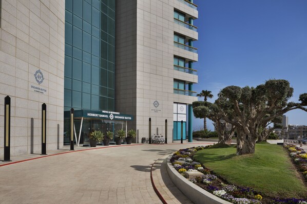 A hotel entrance with a modern facade, a landscaped garden, and a prominent tree.