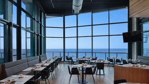 Un restaurante moderno con vistas panorámicas al mar, que ofrece una combinación de asientos tipo cabina y mesas, además de una zona de bar central.