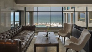 Un área de estar moderna con un sofá de cuero, una mesa de centro de madera y dos sillones. Un ventanal ofrece vistas al océano y a una piscina.